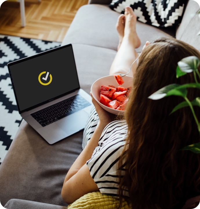 Femme allongée sur un canapé, mangeant un bol de fruits, avec un ordinateur portable à côté d’elle affichant le logo Norton